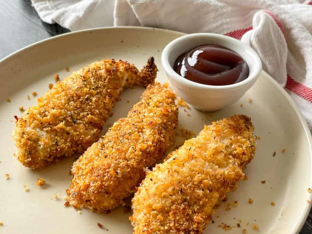 Three breaded and baked chicken tenders on a plate with a small bowl of dipping sauce.