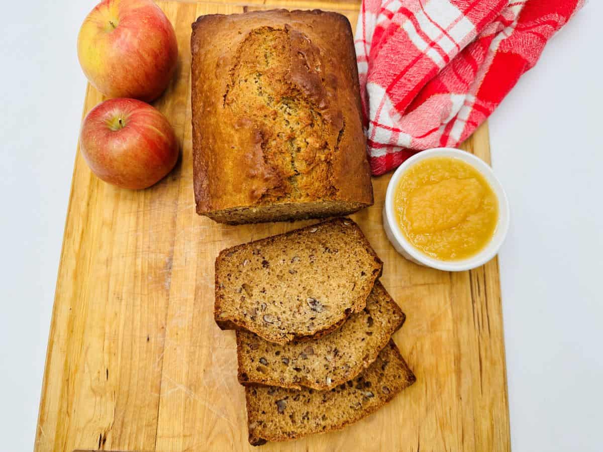 A loaf of applesauce bread with three slices cut, two apples, a bowl of applesauce, and a red and white checkered cloth on a wooden cutting board.