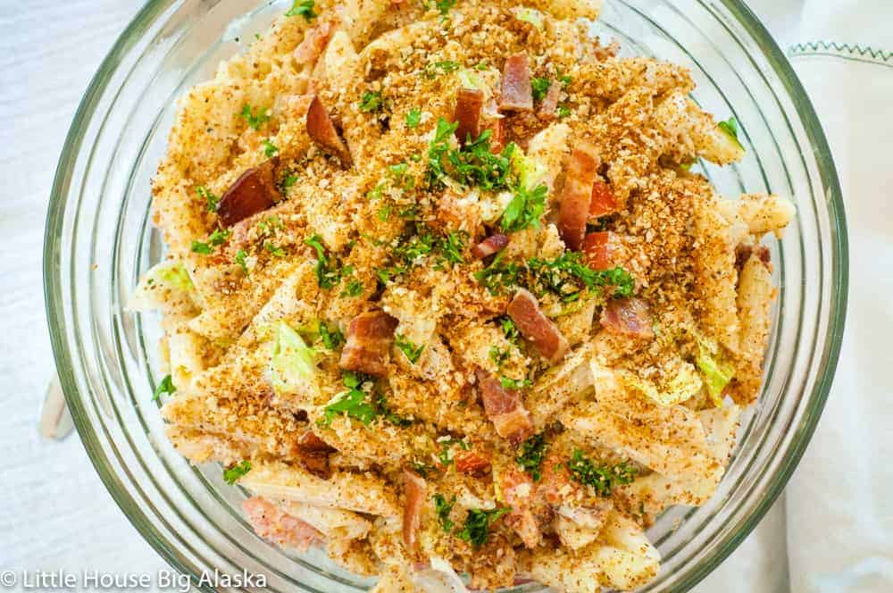 A bowl of pasta salad topped with breadcrumbs, bacon bits, and chopped herbs, placed on a white tablecloth.