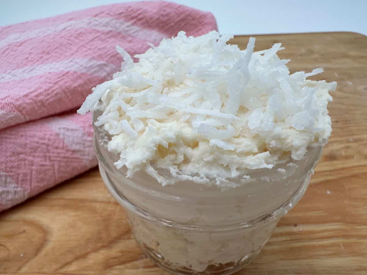 A glass jar filled with a white creamy substance topped with shredded coconut sits on a wooden surface. A pink cloth is in the background.