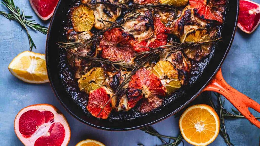 A baked dish garnished with slices of citrus fruits and topped with sprigs of rosemary, surrounded by halved grapefruit, lemon, and orange on a blue surface.