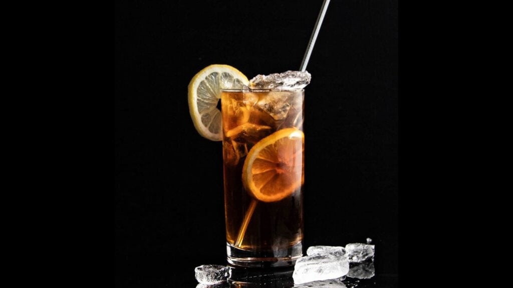 A tall glass of iced tea garnished with lemon slices and filled with ice cubes, with a metal straw inserted, set against a black background.