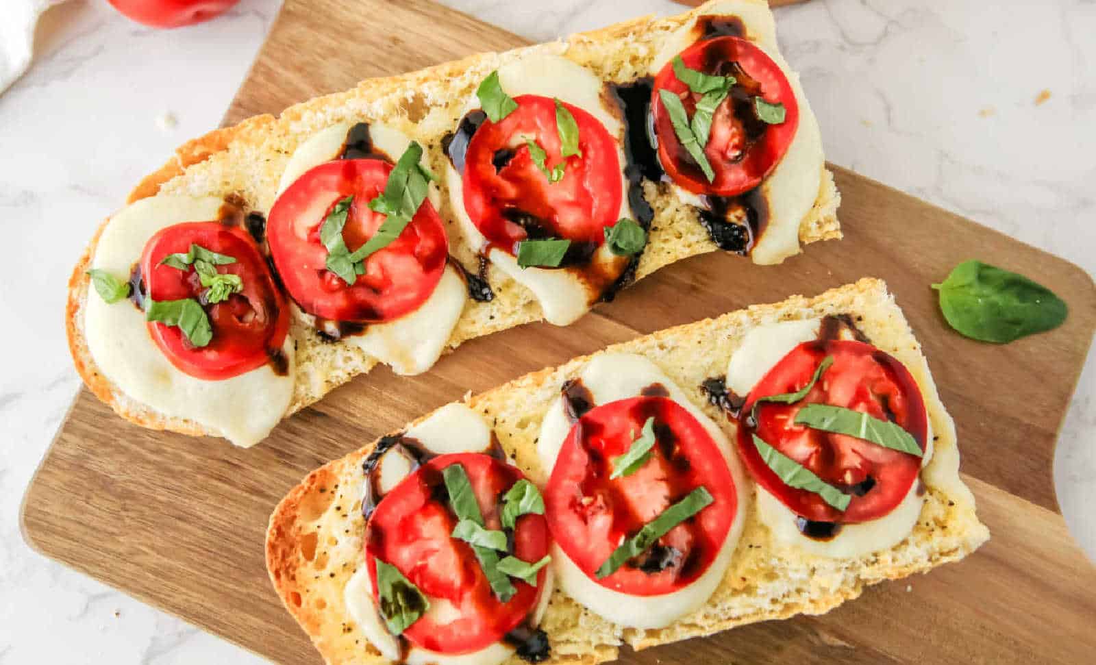 Slices of bread topped with melted cheese, tomato slices, basil leaves, and balsamic glaze, arranged on a wooden board.