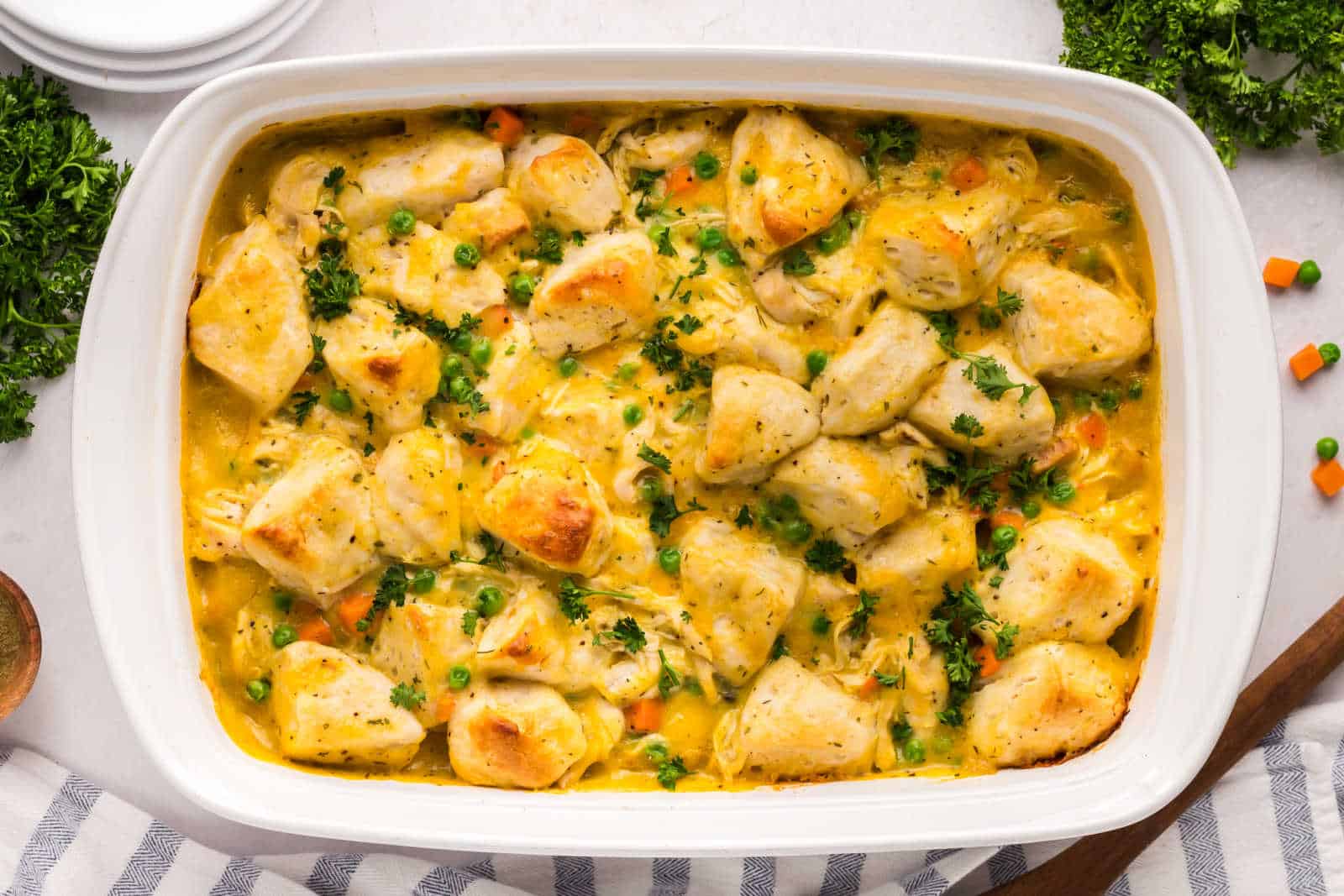 A casserole dish filled with baked chicken, mixed vegetables, and creamy sauce, with a wooden spoon resting inside. 