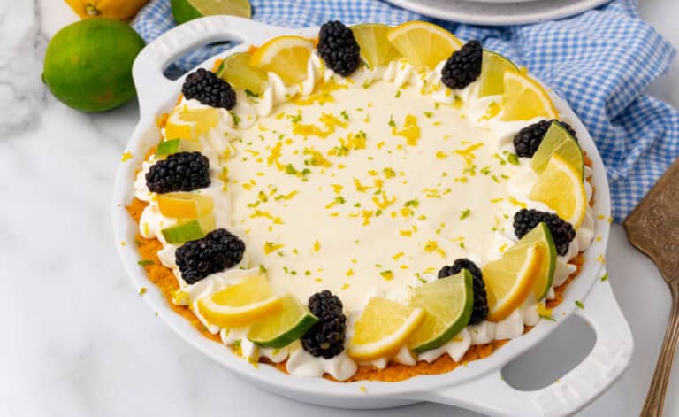 A pie with a creamy filling is garnished with slices of lime and orange, blackberries, and whipped cream, placed on a checkered blue cloth.