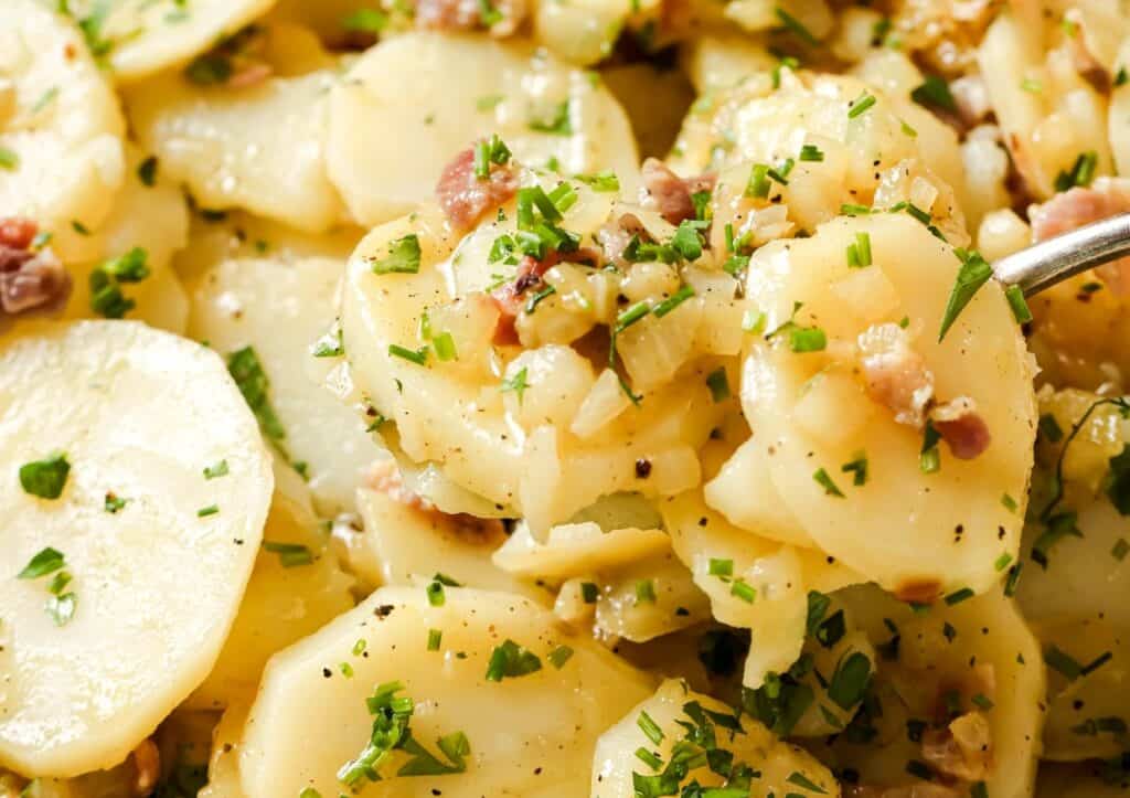 Close-up of a plate of potato salad garnished with chopped parsley and bits of bacon, with a metal spoon serving a portion.