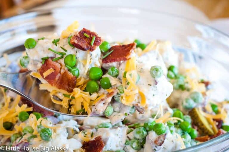 A spoon scooping a potato salad mixed with peas, bacon bits, and shredded cheddar cheese from a clear glass bowl.