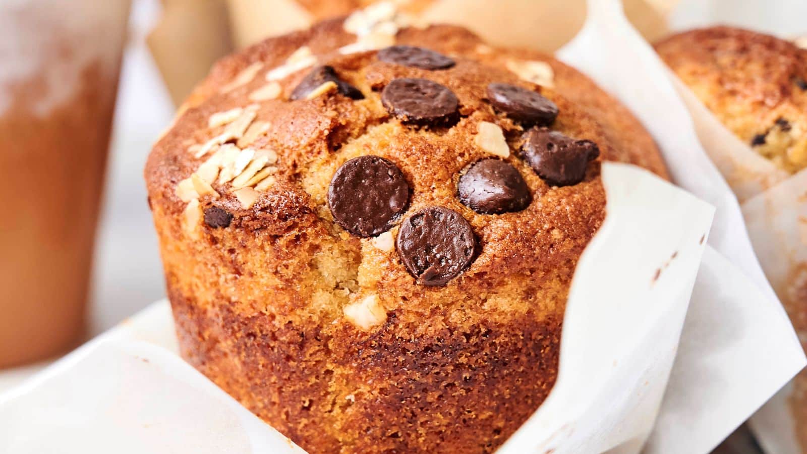 Close-up of a freshly baked muffin topped with chocolate chips and oats, wrapped in a paper liner. A blurred drink and other baked goods are visible in the background.