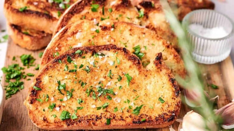 Slices of toasted garlic bread garnished with parsley and sea salt, arranged on a wooden cutting board with a small bowl of salt in the background.