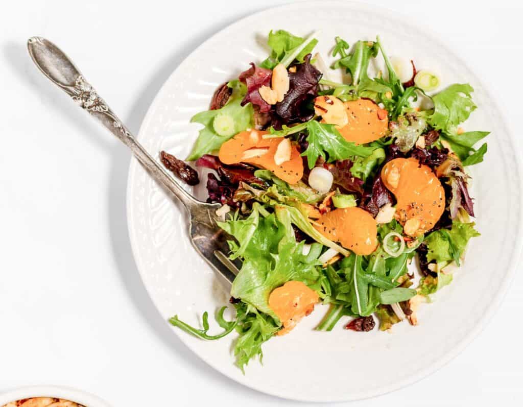 A white plate with a mixed green salad, topped with mandarin orange slices, sliced almonds, and raisins, with a fork on the side.