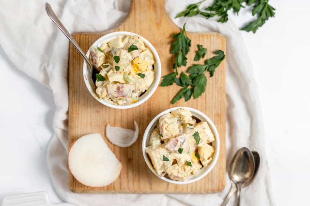 Two bowls of potato salad sit on a wooden cutting board, garnished with chopped parsley. A white onion, spoon, and parsley are also on the board. A white cloth is draped in the background.