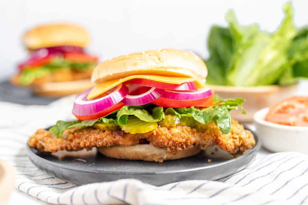 A crispy chicken sandwich with red onion, lettuce, cheddar cheese, and a bun, placed on a dark plate with a sandwich and fresh lettuce in the background.