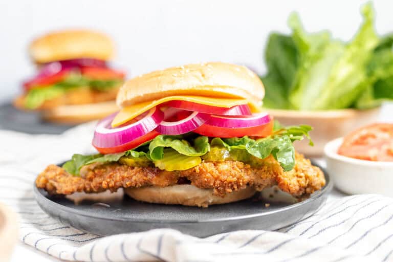 A crispy chicken sandwich with red onion, lettuce, cheddar cheese, and a bun, placed on a dark plate with a sandwich and fresh lettuce in the background.