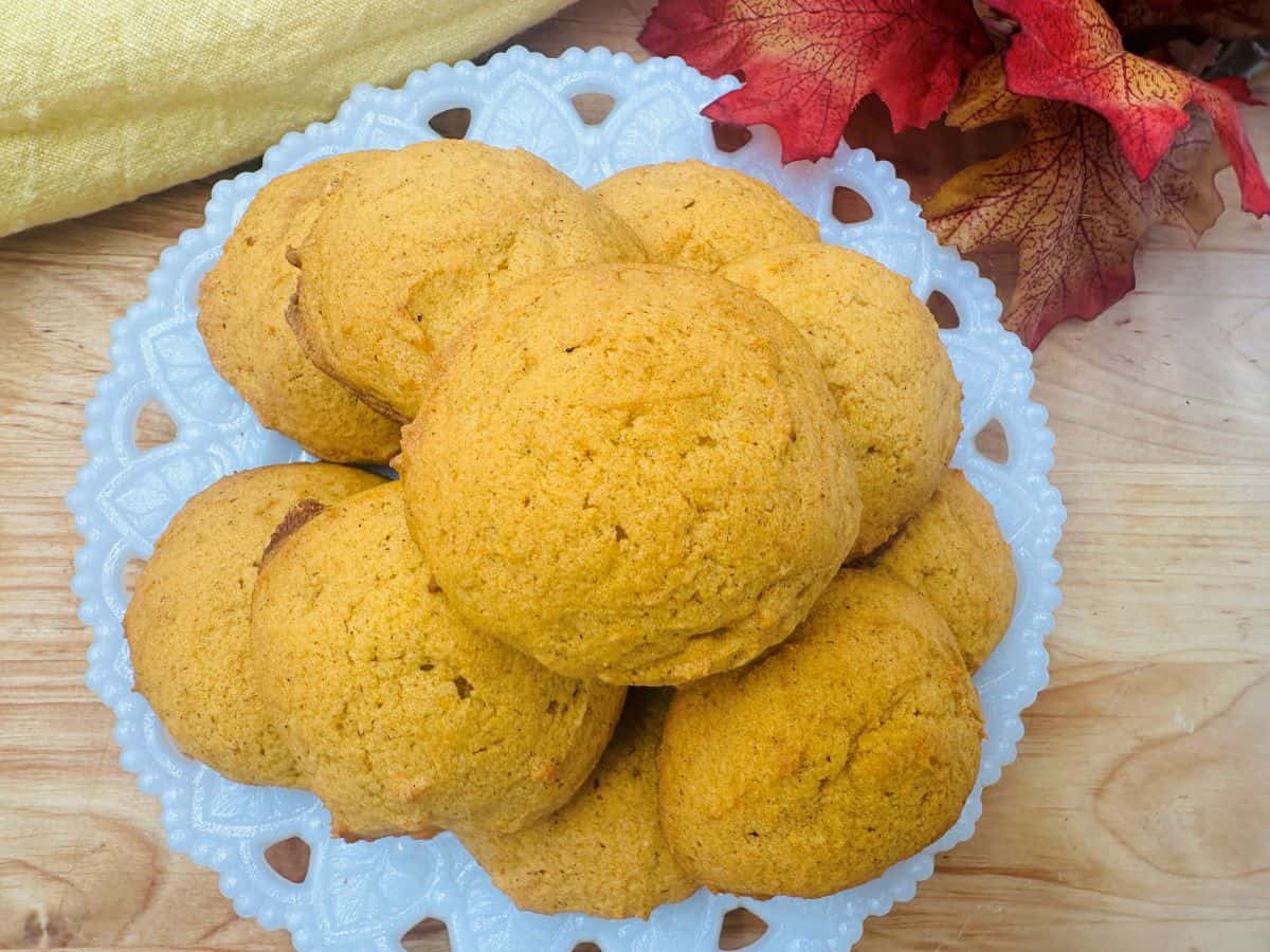 A white plate holds a stack of pumpkin cookies on a wooden table, surrounded by a yellow cloth and autumn leaves.