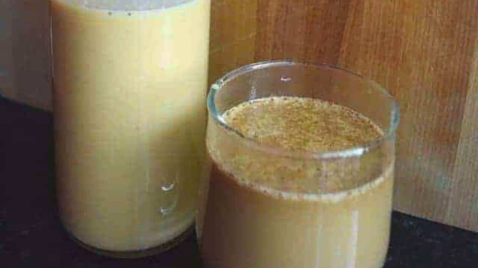 Image shows A glass and a bottle filled with a pumpkin spice latte are placed on a dark surface against a wooden backdrop.