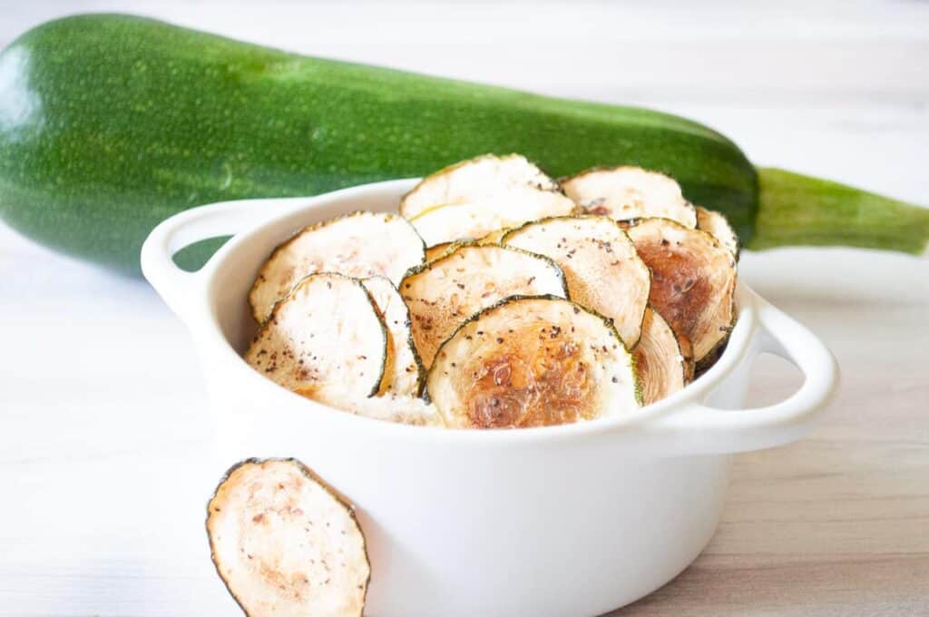 A white bowl filled with baked zucchini chips is placed in front of a whole zucchini on a light-colored surface.