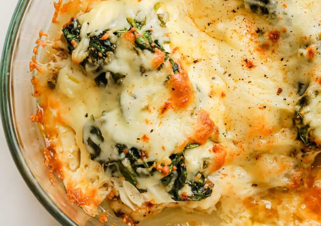 A close-up of a baked dish with melted cheese, spinach, and chicken in a glass baking dish, showing a golden brown crust.