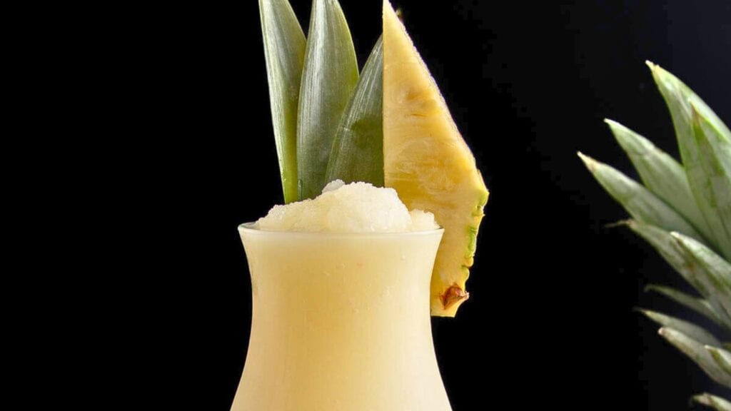 A glass of blended pineapple smoothie garnished with pineapple leaves and a pineapple wedge against a black background.