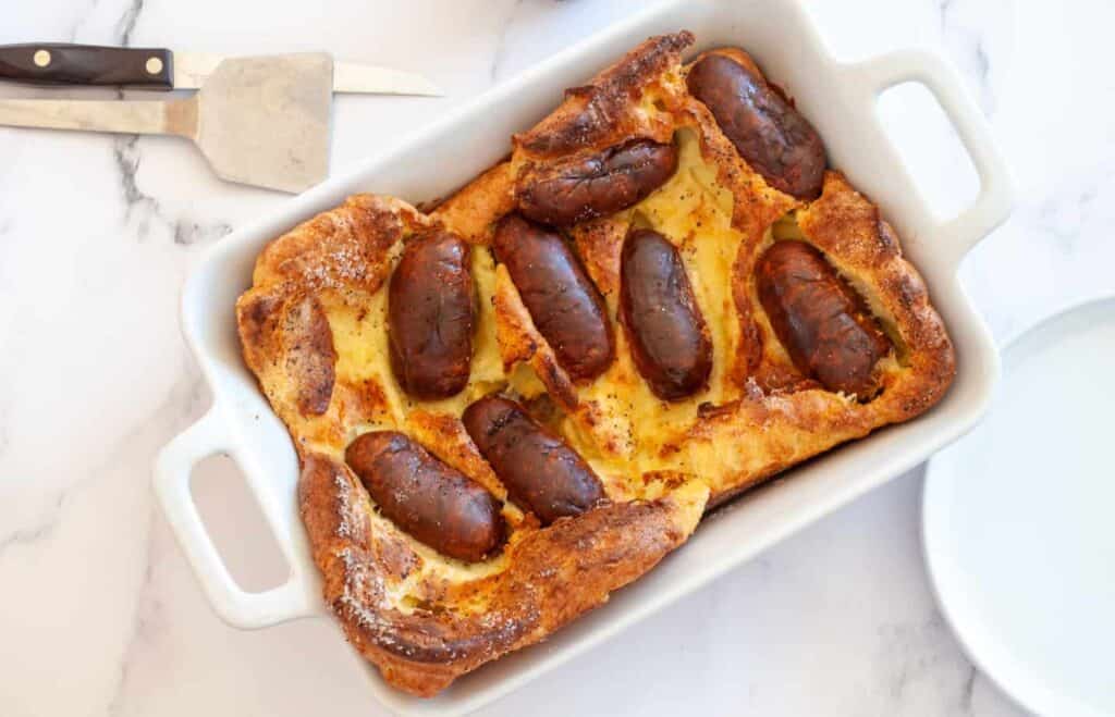 A baked dish featuring sausages embedded in a Yorkshire pudding, placed in a rectangular baking dish. A knife and a serving utensil are nearby on a marble countertop.