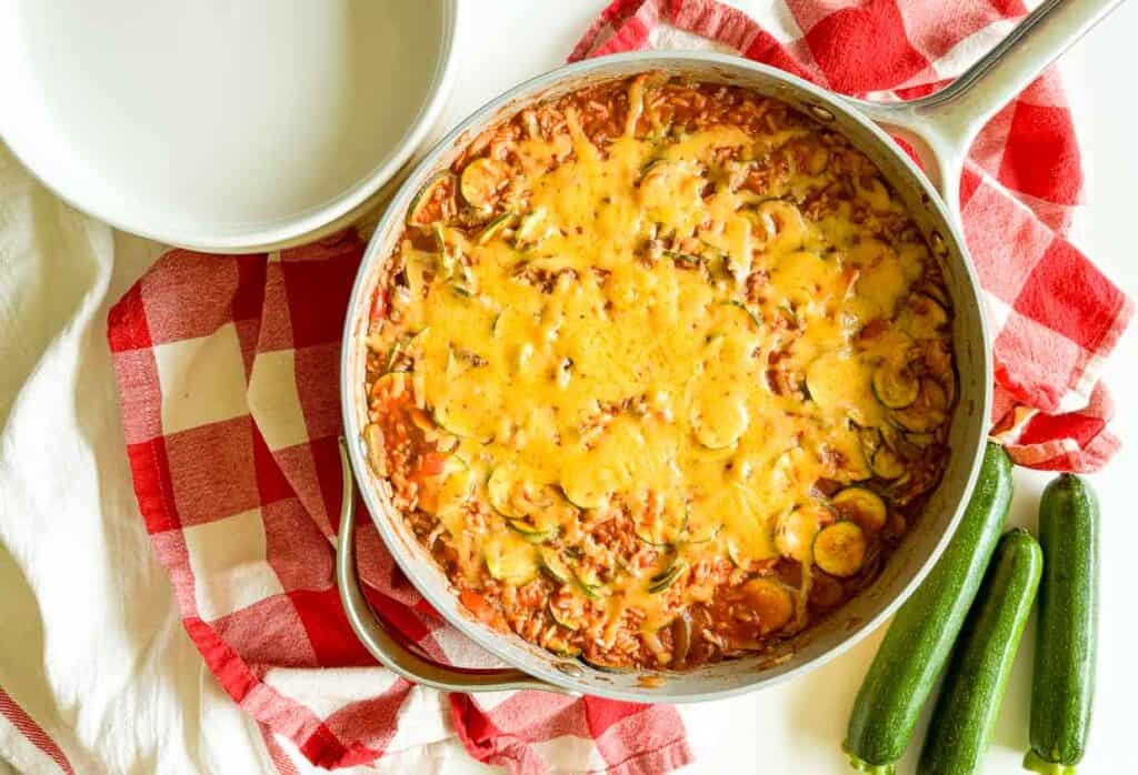 A skillet filled with a cheesy zucchini and ground meat casserole on a red and white checkered cloth, next to three zucchinis and an empty white bowl.