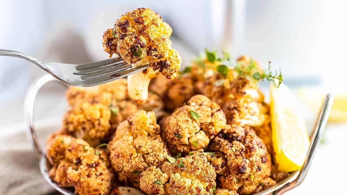 A fork holding a piece of roasted cauliflower garnished with herbs, above a bowl filled with more roasted cauliflower and lemon wedges.