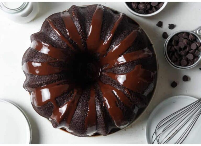 A chocolate bundt cake drizzled with chocolate glaze is surrounded by chocolate chips, a whisk, plates, and a jar of milk.