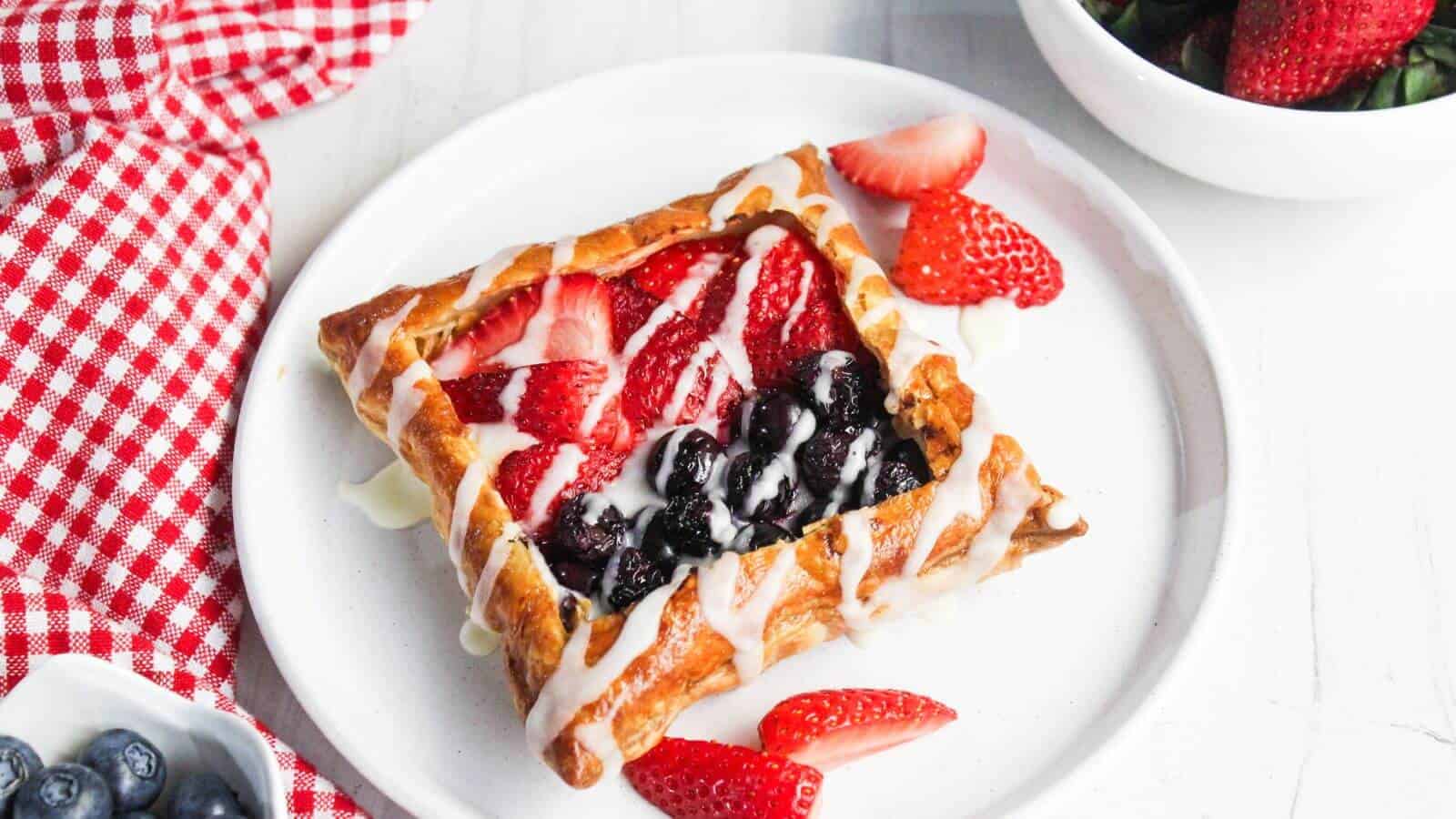 A pastry with berry filling and white drizzle on a white plate is accompanied by strawberry slices. A red and white checkered cloth, blueberries, and a bowl of strawberries are nearby.