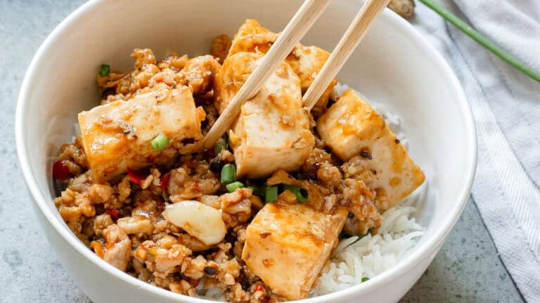 A bowl of rice topped with stir-fried tofu cubes and minced vegetables, garnished with green onions. Chopsticks rest on top.