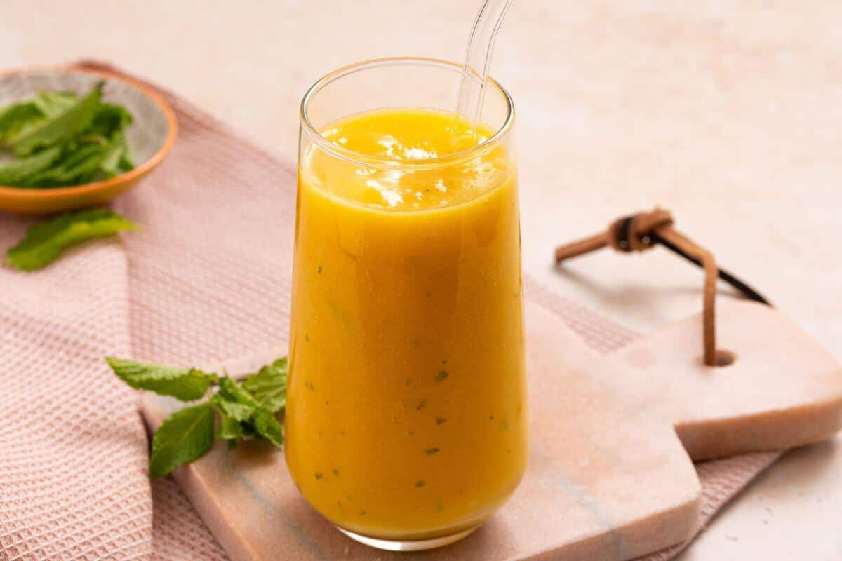 A tall glass of yellow smoothie is placed on a light-colored stone board with a glass straw. Fresh mint leaves and a cloth napkin are next to it.