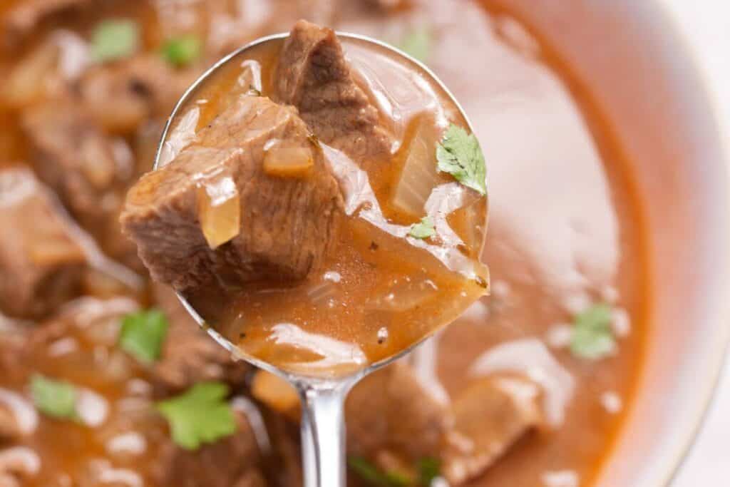 A close-up of a spoonful of beef stew with chunks of beef and vegetables, served in a rich, brown broth with garnished cilantro.