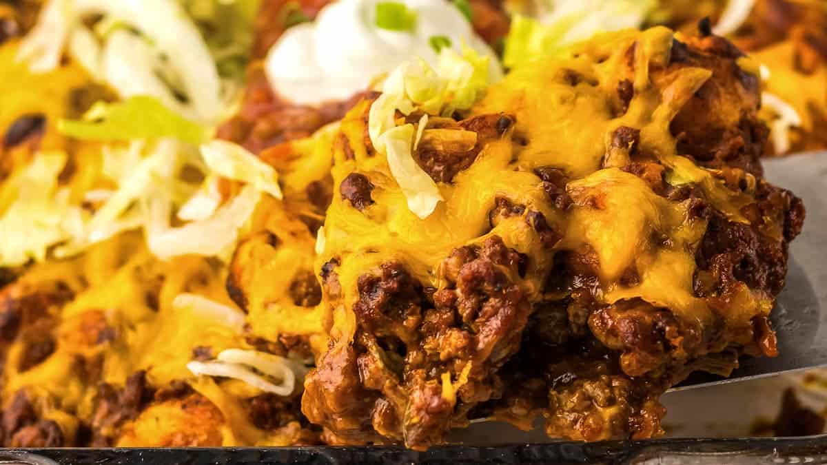 A close-up of a cheesy taco casserole topped with shredded lettuce and sour cream, being served.