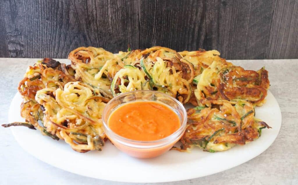 A white plate with crispy vegetable fritters and a small bowl of orange dipping sauce in the center. The background is a dark wood panel.