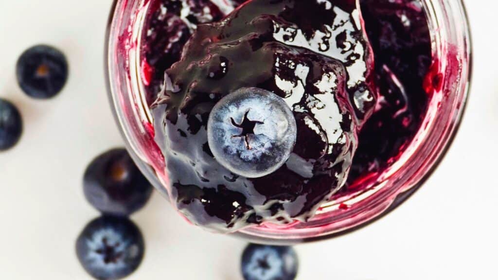 Close-up of a jar of blueberry jam with a single fresh blueberry resting on top, surrounded by a few scattered blueberries.