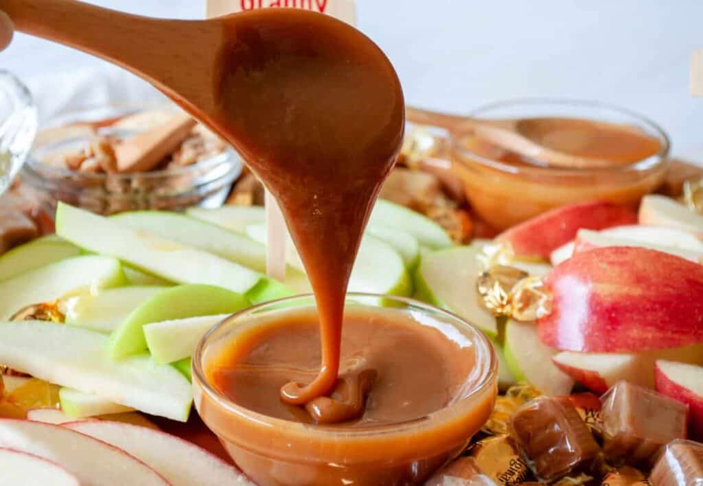 A wooden spoon drizzles caramel into a small bowl. Sliced green and red apples, candy, and a dish of nuts are visible in the background.