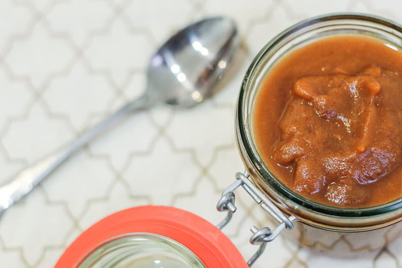 A jar of thick brown sauce with visible chunks sits on a patterned surface next to a silver spoon.