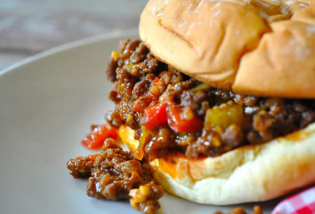 A close-up image of a sloppy joe sandwich on a plate with ground beef and diced vegetables spilling out of a bun.