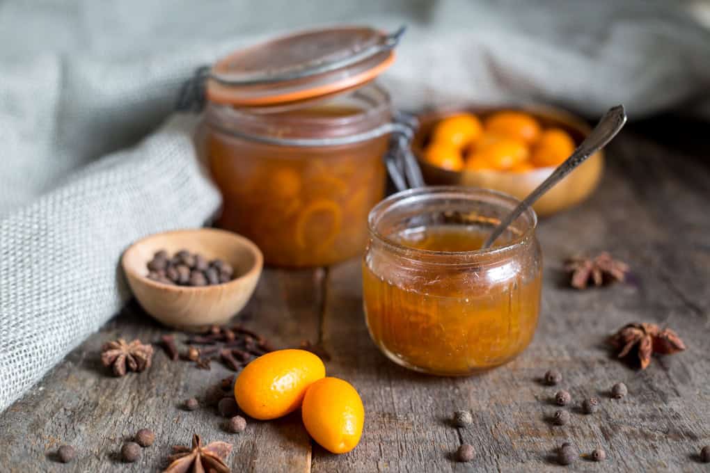 A jar of orange marmalade with a spoon inside sits on a wooden surface next to a larger sealed jar, three kumquats, a small bowl of spices, and scattered spice seeds and star anise around.