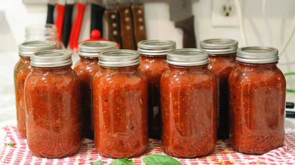 Eight jars filled with homemade tomato sauce are lined up on a kitchen counter covered with a red and white towel. Knives and kitchen tools are visible in the background.