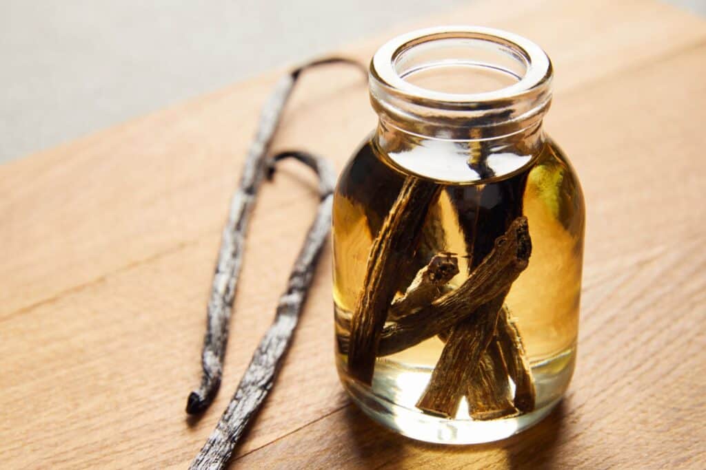 A small glass bottle filled with a yellow liquid and several vanilla beans inside, placed on a wooden surface with additional vanilla pods next to it.