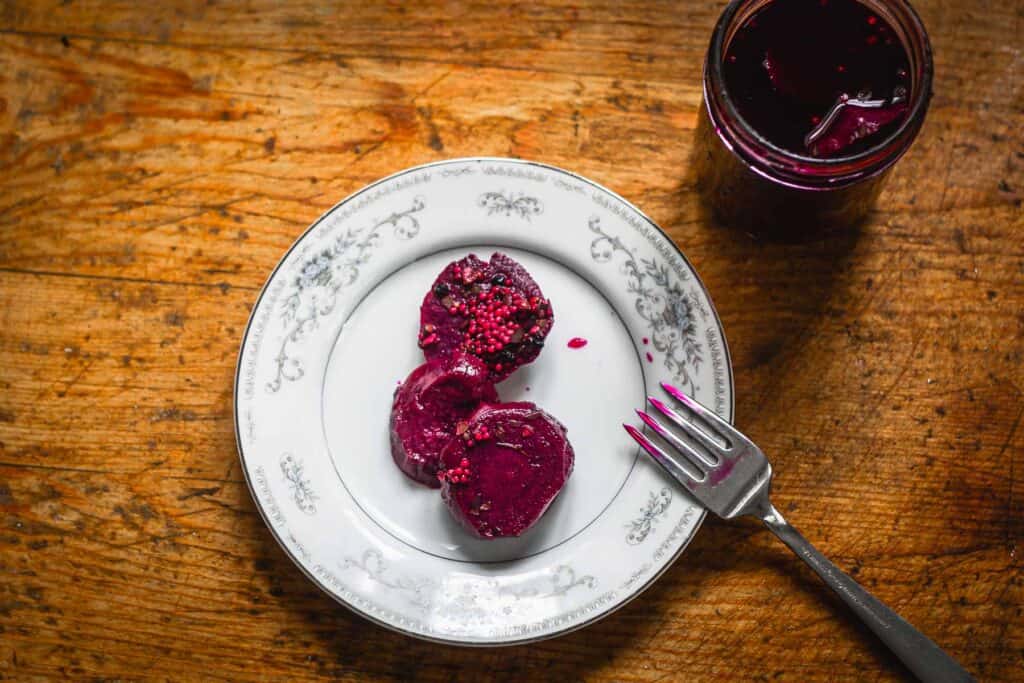 A plate with three slices of pickled beetroot and a fork next to a jar of pickled beets on a wooden table.