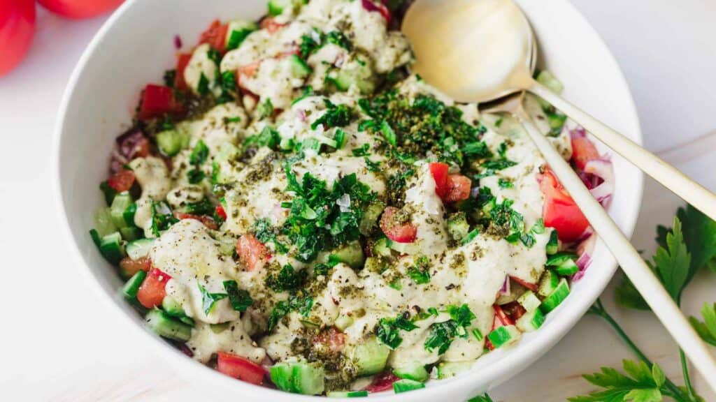 A dish filled with a fresh salad consisting of chopped tomatoes, cucumbers, and herbs, topped with a creamy dressing and garnished with ground black pepper and chopped greens, with two spoons placed inside.