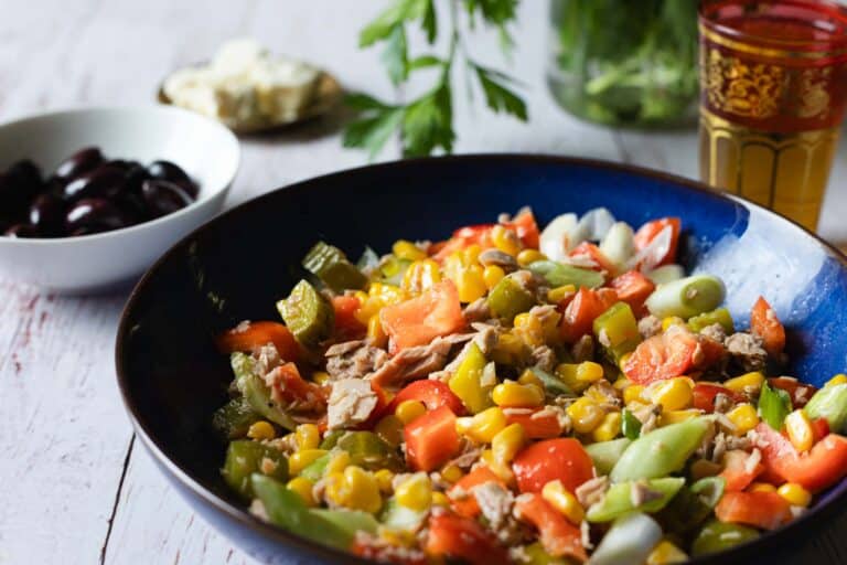 A colorful vegetable salad with corn, bell peppers, and green onions in a blue bowl, accompanied by a bowl of black olives, a piece of cheese, and a glass of drink on a wooden table.