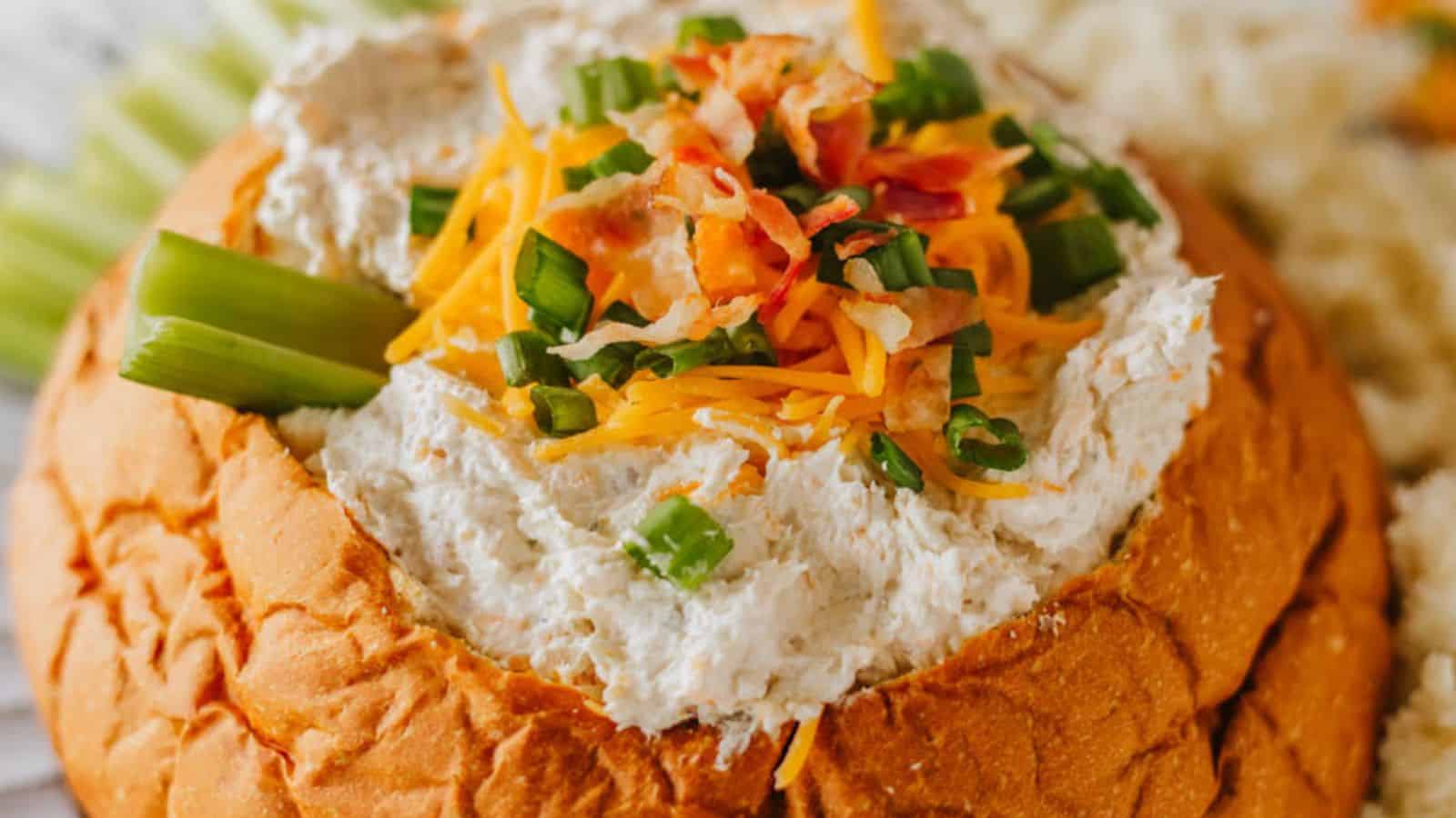 A bread bowl filled with a creamy dip, garnished with shredded cheese, chopped green onions, and bacon bits. Celery sticks are placed on the side.