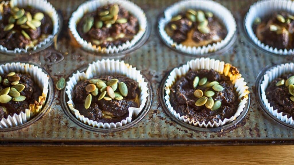 Close-up of six muffins in a metal baking tray, each topped with pumpkin seeds, placed in white paper liners.