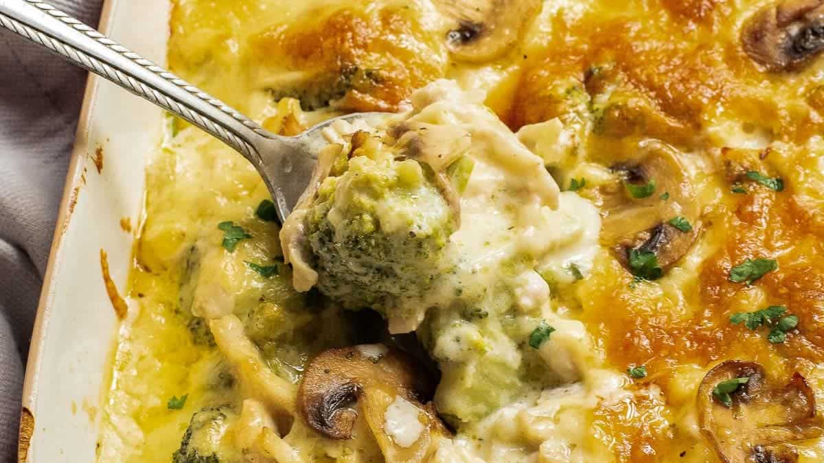 A close-up of a creamy baked casserole with golden-brown cheese, mushrooms, broccoli in a red dish.