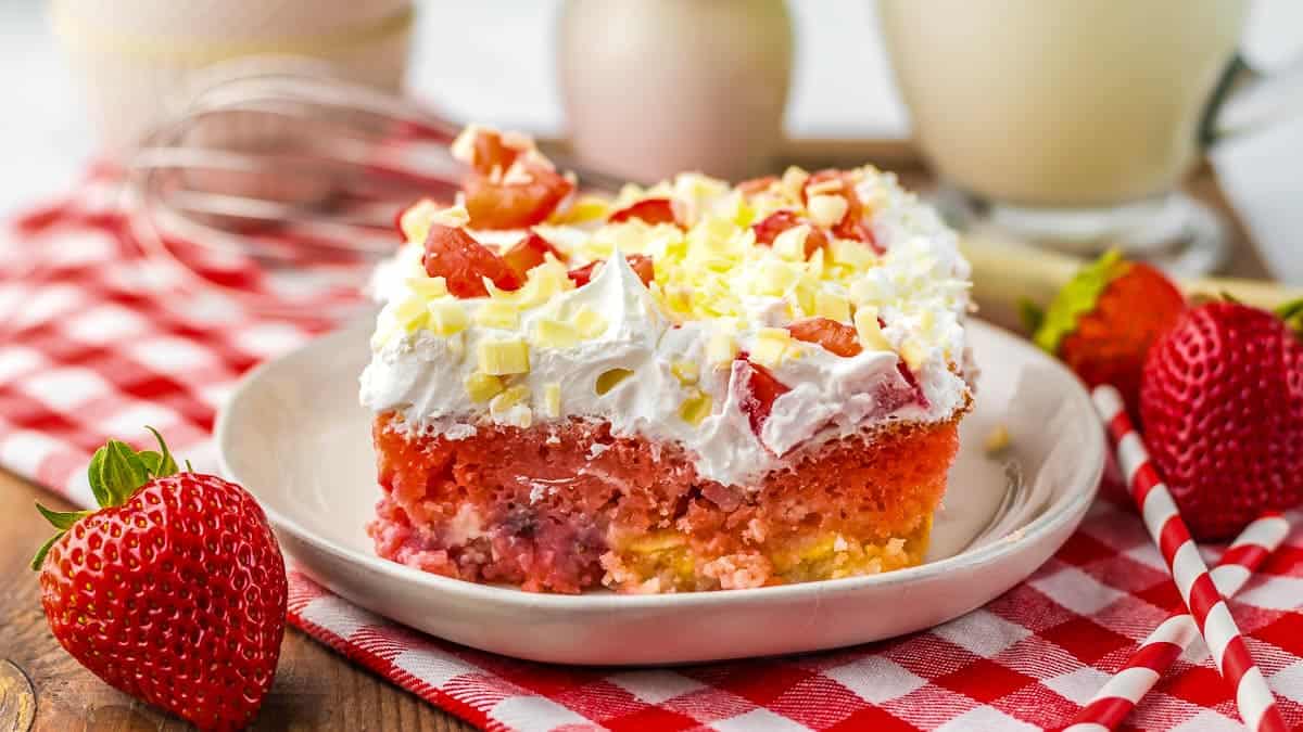 A slice of strawberry poke cake with white frosting and white chocolate shavings on a white plate. The plate is placed on a red and white checkered cloth.