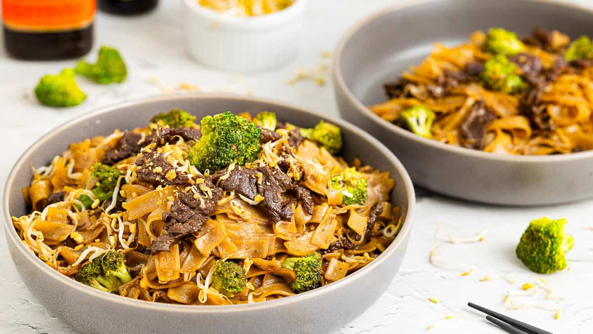 Two bowls of beef chow fun stir-fried with broccoli and bean sprouts, garnished with sesame seeds. The dishes are served in gray bowls, and the background shows another bowl with ingredients. A pair of chopsticks is placed beside the front bowl.