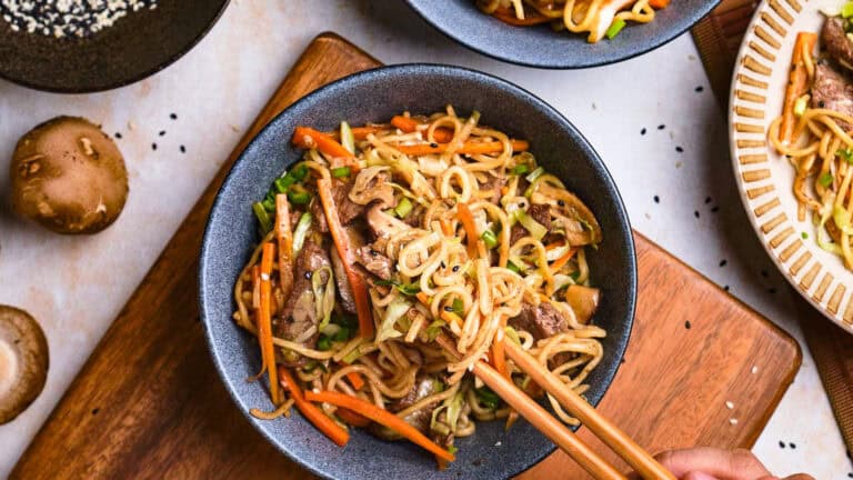 A bowl of stir-fried noodles with vegetables and meat, garnished with black sesame seeds. Chopsticks are picking up a portion. Mushrooms and a plate are visible nearby.