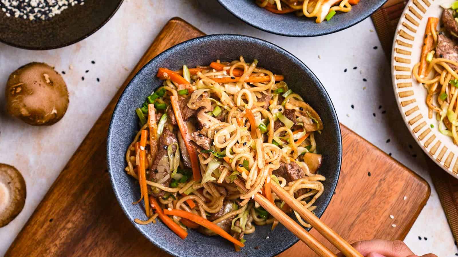 A close-up of a bowl of stir-fried noodles with vegetables and slices of meat, garnished with sesame seeds. A hand is using chopsticks to pick up the noodles. Other bowls and a cutting board are partially visible in the background.