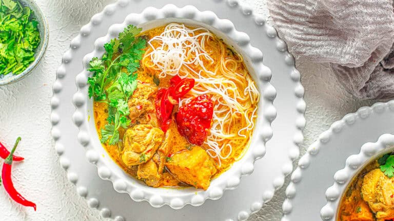 A bowl of curry soup with noodles, chicken pieces, red chili slices, a dollop of red sauce, and cilantro garnish on a textured white surface.